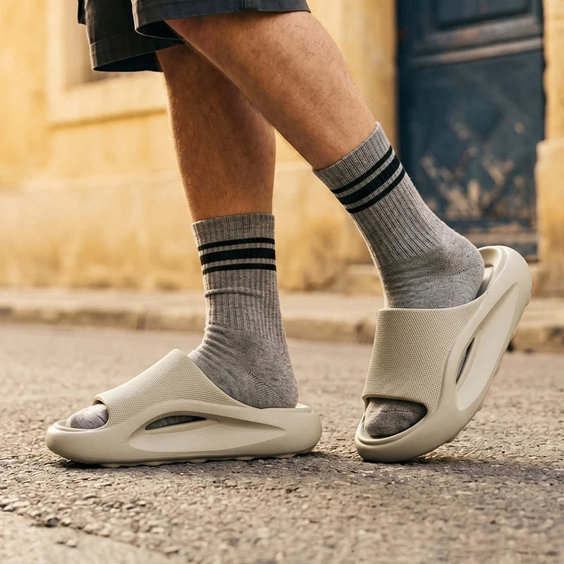 A person wearing gray crew socks with black stripes and off-white molded slide sandals on a street, with a blurred beige building and dark door in the background.